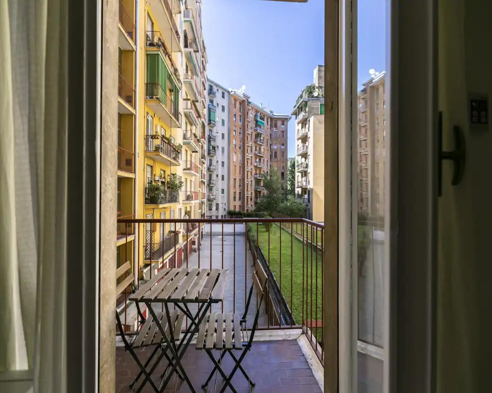 Pequeño balcón amueblado con mesa y dos sillas que da a un patio interior y edificios vecinos. Vista urbana luminosa y acogedora visible a través de la puerta abierta.