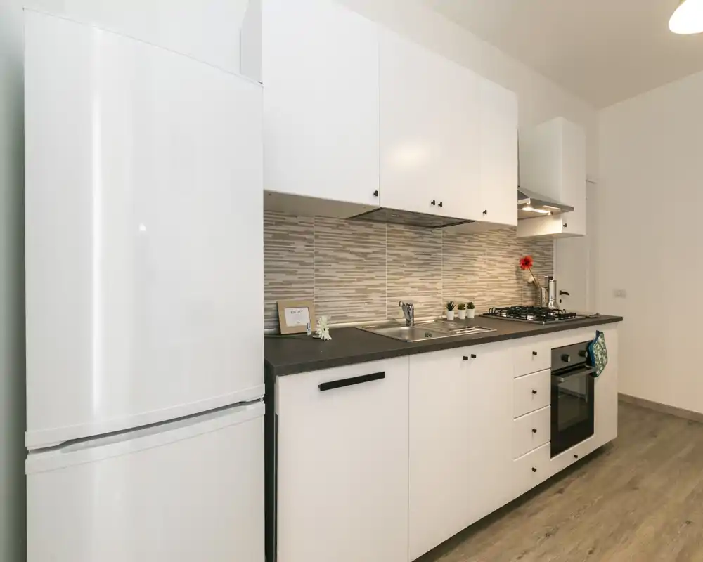 Cocina moderna y compacta con armarios blancos, horno y frigorífico integrados, salpicadero de azulejos y encimera ordenada con pequeños detalles decorativos.