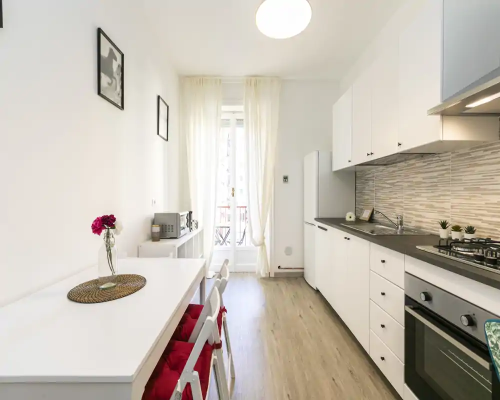 Cocina moderna y luminosa con muebles blancos, horno integrado y mesa de comedor. La luz natural entra por la puerta acristalada que da a un pequeño balcón.