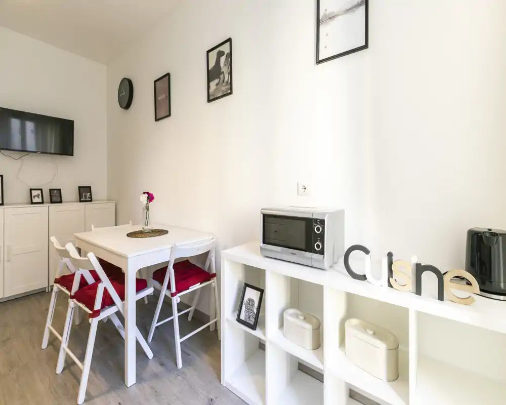 Rincón de comedor moderno y luminoso con pequeña mesa blanca, sillas plegables con cojines rojos y estantería abierta con microondas—ideal para apartamentos compactos.