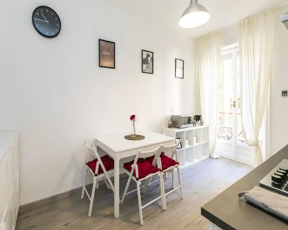 Zona de comedor luminosa y minimalista con mesa blanca, cuatro sillas con cojines rojos y luz natural procedente de las puertas que dan a un pequeño balcón.
