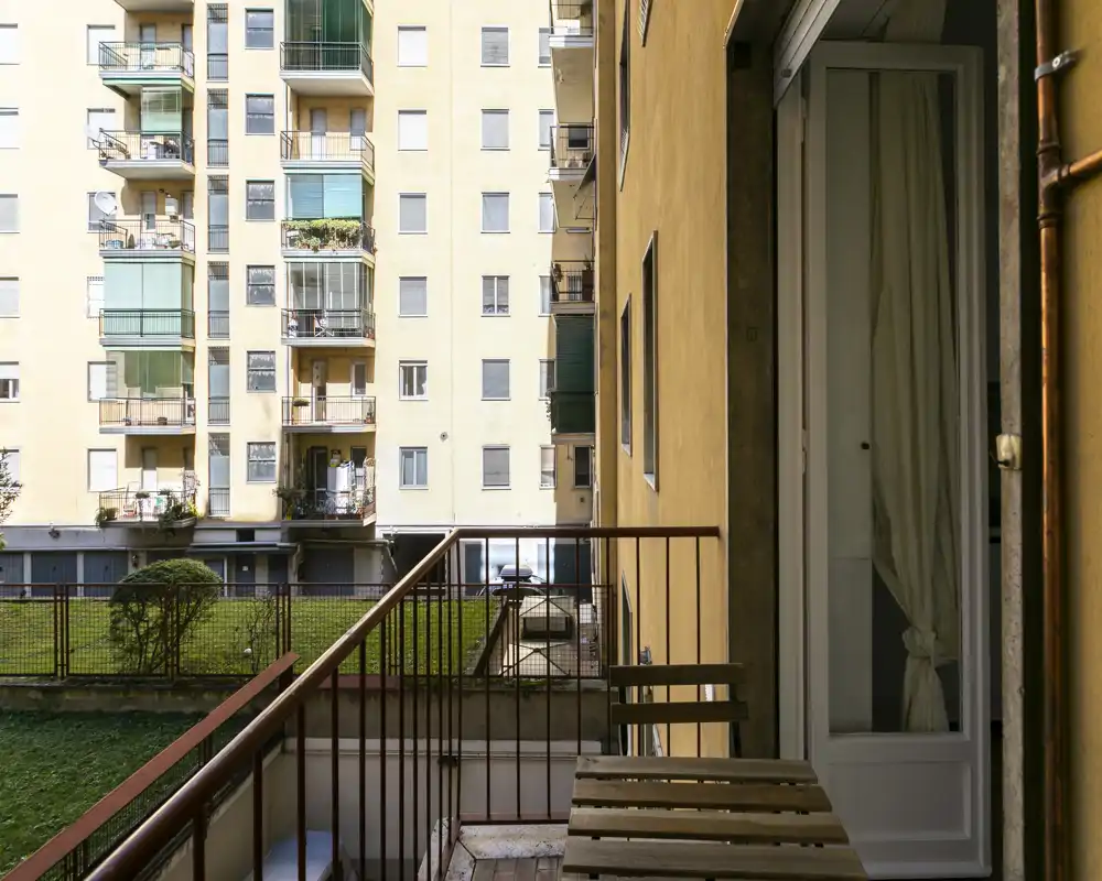 Pequeño balcón privado con vistas a un patio interior y las fachadas de los edificios vecinos; mesa plegable de madera y barandilla visibles, ideal para un café por la mañana.