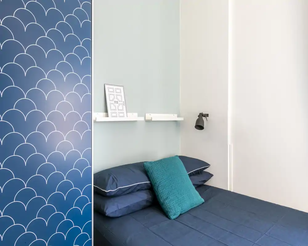 Dormitorio compacto y moderno con cama azul marino, cojín turquesa y un panel de armario decorado. Estanterías sencillas y una lámpara de pared crean un rincón de descanso ordenado y con estilo.