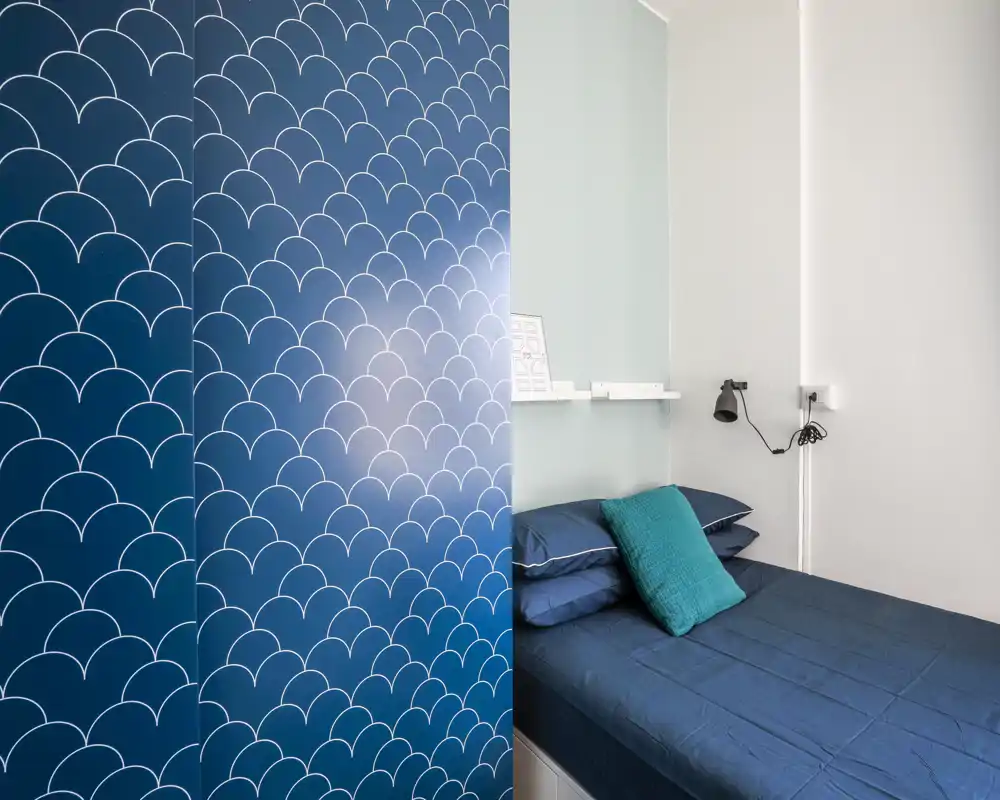 Dormitorio pequeño con cama azul bien hecha, cojín turquesa y biombó decorativo; estilo moderno y luz suave.