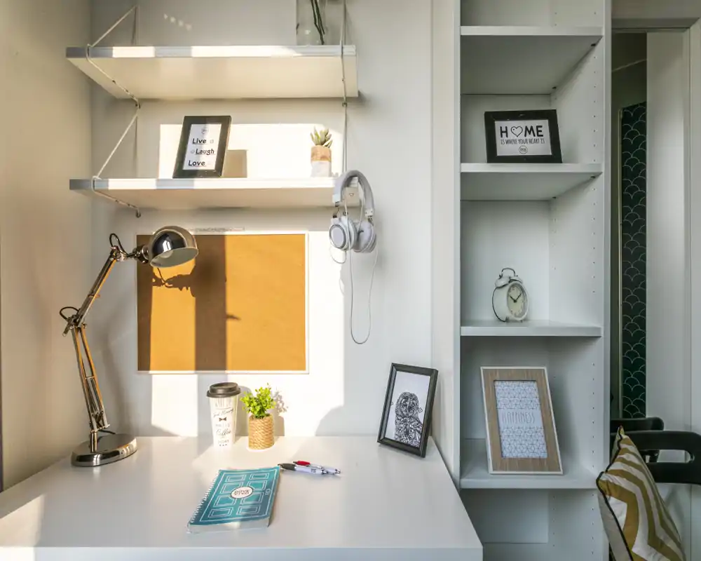 Rincón de oficina en casa luminoso y ordenado con escritorio blanco, lámpara ajustable, marcos, estanterías y planta decorativa — ideal para trabajar o estudiar.