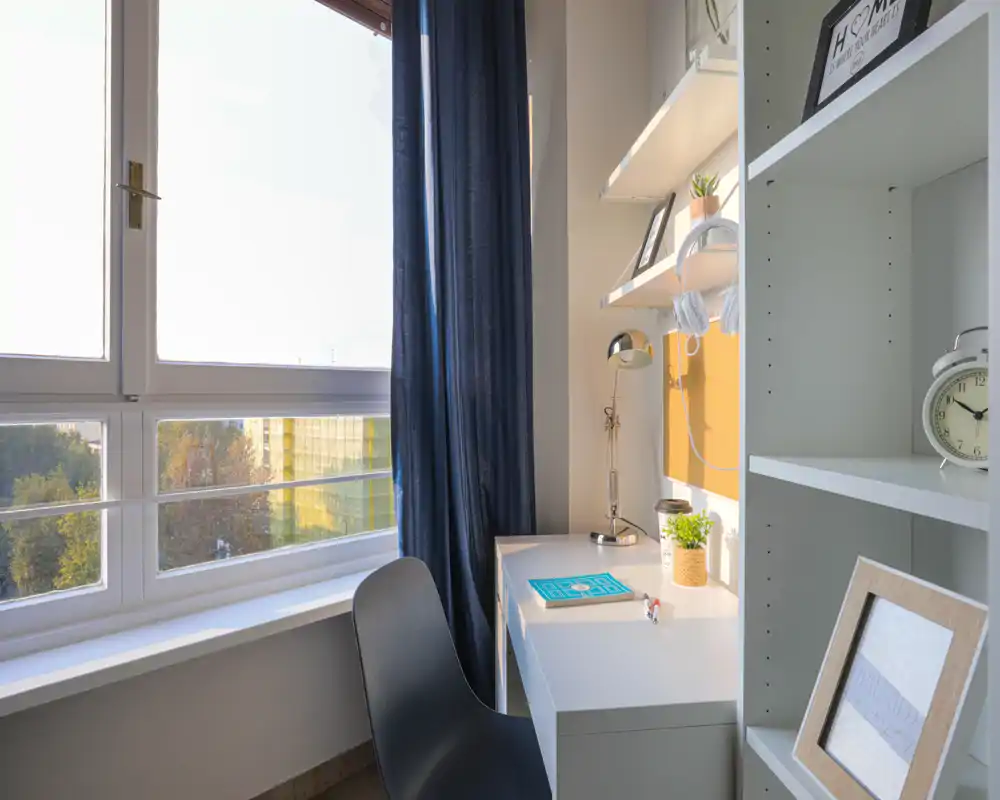 Rincón de trabajo luminoso junto a una gran ventana con mesa blanca, lámpara de estudio, estanterías y elementos decorativos — ideal para trabajar o estudiar con abundante luz natural.