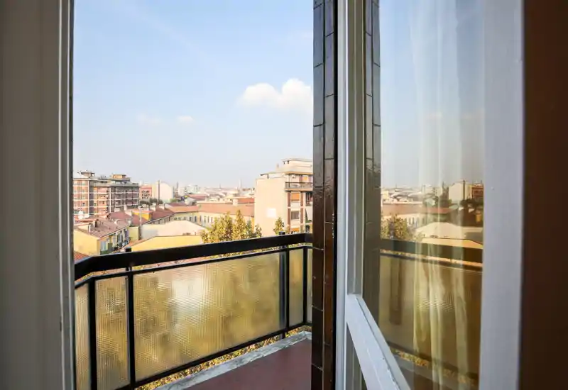 Balcone privato soleggiato con ringhiera in metallo e pannelli di vetro strutturato, con vista chiara sui tetti e gli edifici della città — perfetto per la colazione.
