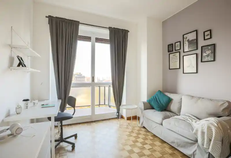 Soggiorno luminoso e ben arredato con divano confortevole, quadri decorativi e un angolo lavoro vicino alla porta del balcone che illumina l'ambiente con luce naturale.