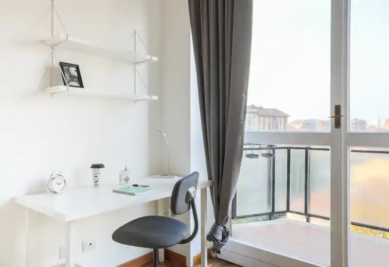 Angolo camera luminoso con scrivania e sedia accanto alla porta del balcone; arredo minimal con tenda pesante e mensole sospese.