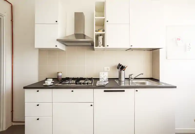 Cucina moderna e compatta con mobili bianchi, piano cottura a gas, lavello in acciaio inox e cappa elegante; luminosa, ordinata e pronta all'uso.