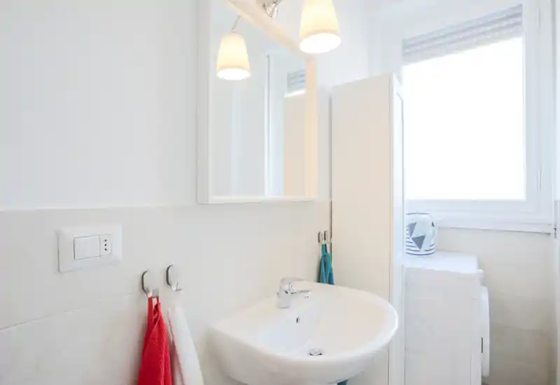 Bagno moderno e luminoso con lavabo a parete, specchio e ganci per gli asciugamani; luce naturale da una finestra ampia e piastrelle bianche pulite.