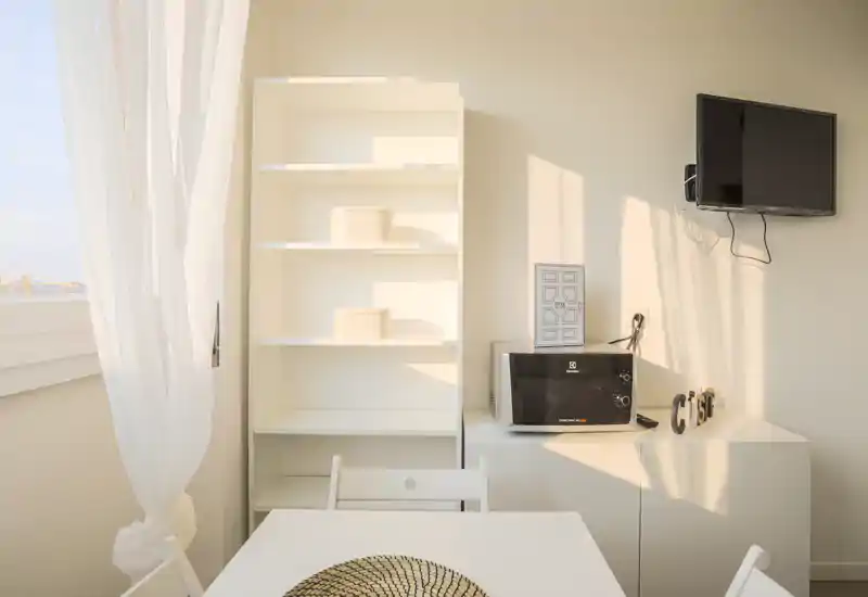 Angolo cucina/pranzo luminoso con un piccolo tavolo, scaffalatura e microonde su un mobile; tende leggere e luce naturale calda creano un'atmosfera minimal e accogliente.