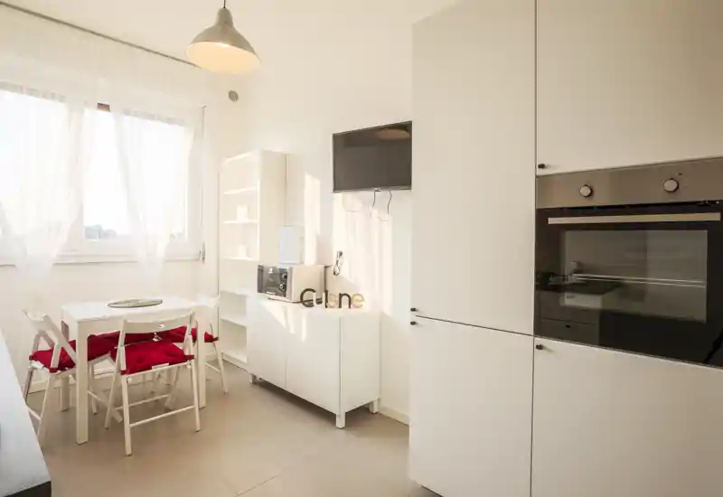 Cucina luminosa e minimal con forno incassato, piccolo tavolo da pranzo e sedie con cuscini rossi; la luce naturale rende l'ambiente arioso e accogliente.