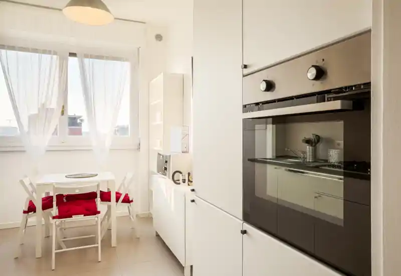 Cucina moderna e luminosa con forno incassato e piccolo angolo pranzo; mobili bianchi e sedie con cuscini rossi rendono lo spazio fresco e accogliente con molta luce naturale.