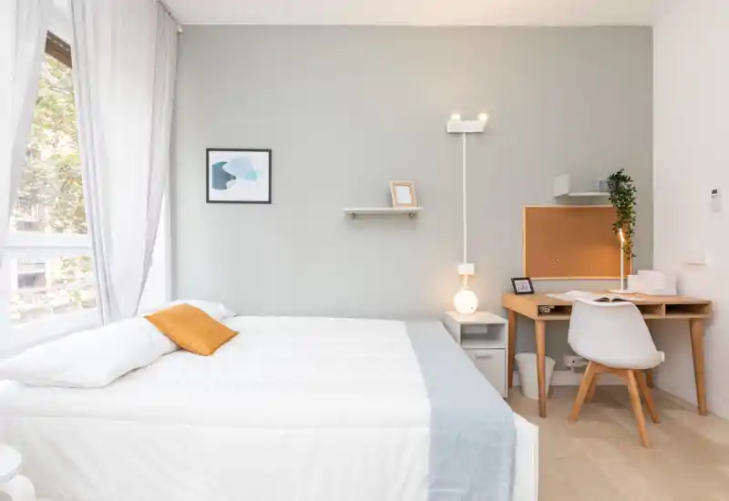 Camera da letto luminosa e minimalista con letto matrimoniale ben rifatto, biancheria chiara e una piccola scrivania in legno: ideale per soggiorni riposanti o un angolo lavoro compatto.