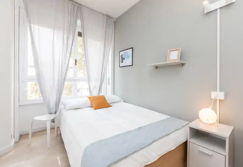 Camera da letto luminosa e moderna con letto matrimoniale ben rifatto, pareti grigio chiaro e ampia finestra che offre luce naturale. Arredo minimal con comodino, lampada e una mensola con cornice.