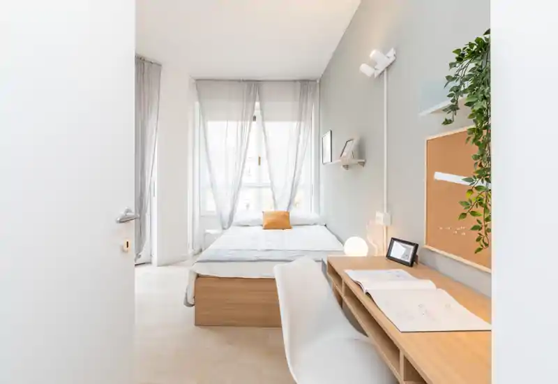 Camera da letto luminosa e minimal con letto matrimoniale al centro sotto tende leggere e una piccola scrivania in legno a parete, ideale per soggiorni riposanti o lavoro da remoto.