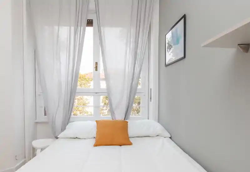 Camera da letto luminosa e minimal con letto bianco, pareti grigio chiaro e un cuscino ocra al centro davanti a una grande finestra che illumina l’ambiente.