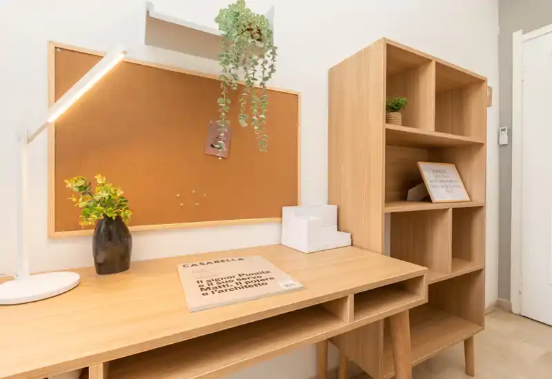 Angolo di lavoro luminoso e minimale con scrivania in legno, bacheca in sughero e scaffalatura coordinata, ideale come home office o studio.