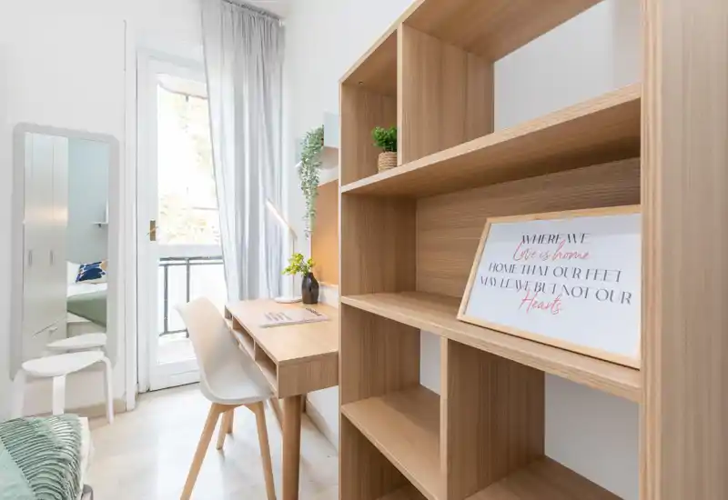 Angolo di camera luminoso e moderno con scrivania e scaffalatura in legno. Luce naturale dalla porta sul balcone, arredi minimali e letto visibile sul lato sinistro.