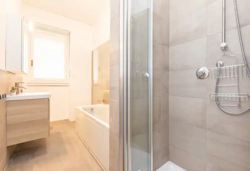 Bagno moderno e luminoso con doccia, vasca e mobile lavabo sospeso. Piastrelle neutre e luce naturale donano un aspetto pulito e contemporaneo.