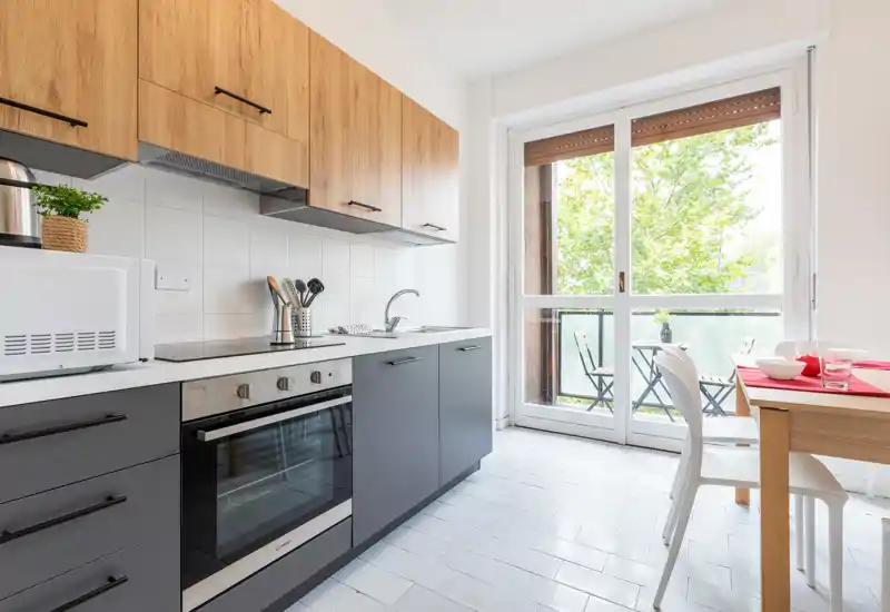 Cucina moderna con pensili in legno e basi scure, forno incorporato e un piccolo tavolo da pranzo accanto a una porta finestra luminosa che dà sul balcone.