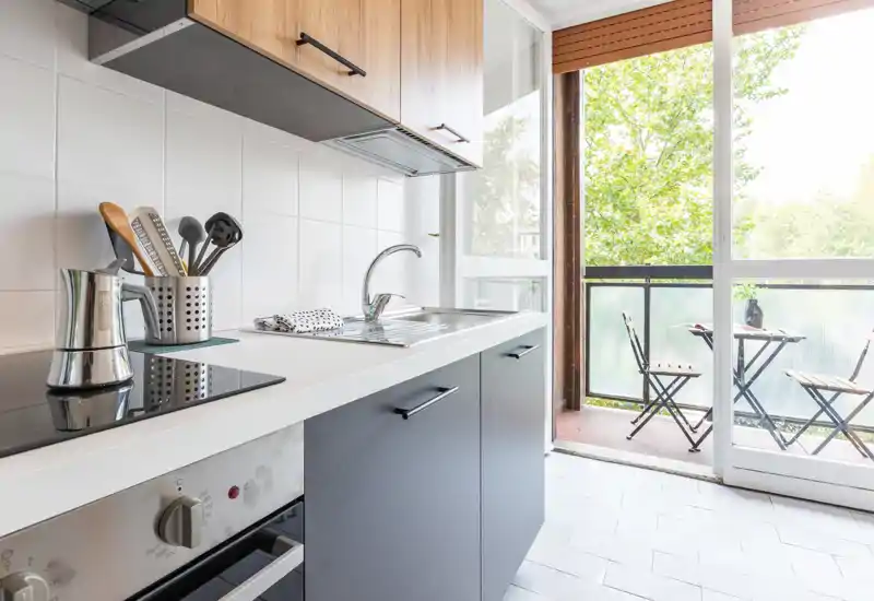 Cucina compatta e moderna con piano bianco, piano cottura a induzione, lavello in acciaio e pensili in legno. Ampia porta scorrevole si apre su un piccolo balcone con zona relax e molta luce naturale.