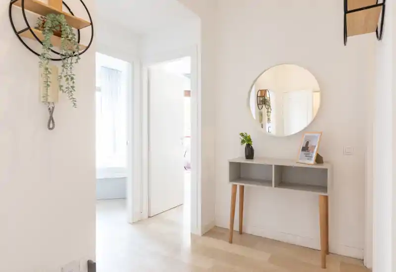 Ingresso luminoso e minimal con un piccolo mobile consolle, specchio rotondo a parete e decorazioni discrete — un'area d'ingresso accogliente e ben illuminata.