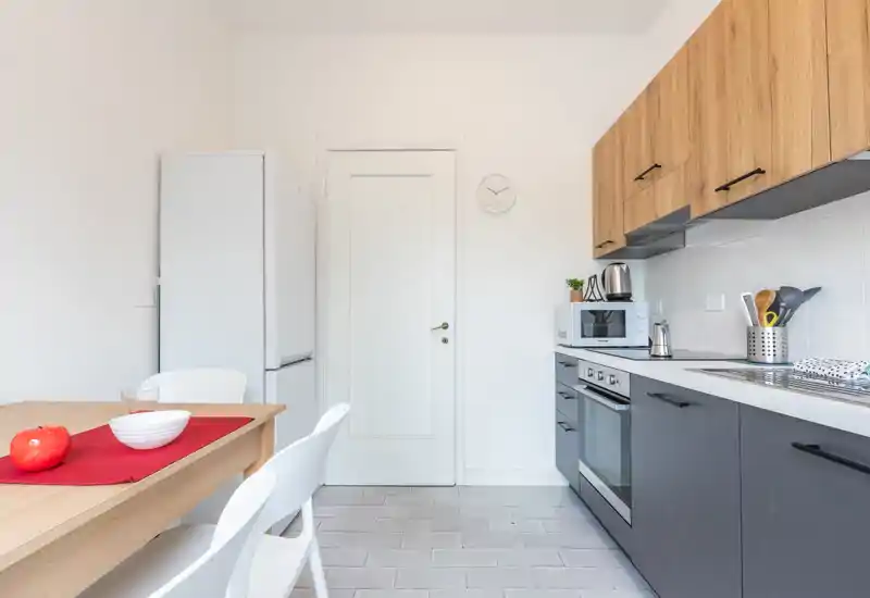 Cucina moderna e compatta con mobili inferiori scuri e pensili in legno, forno integrato, microonde, frigorifero e un piccolo tavolo da pranzo per due persone. Luminoso, pulito e funzionale con piccoli elettrodomestici sul piano.