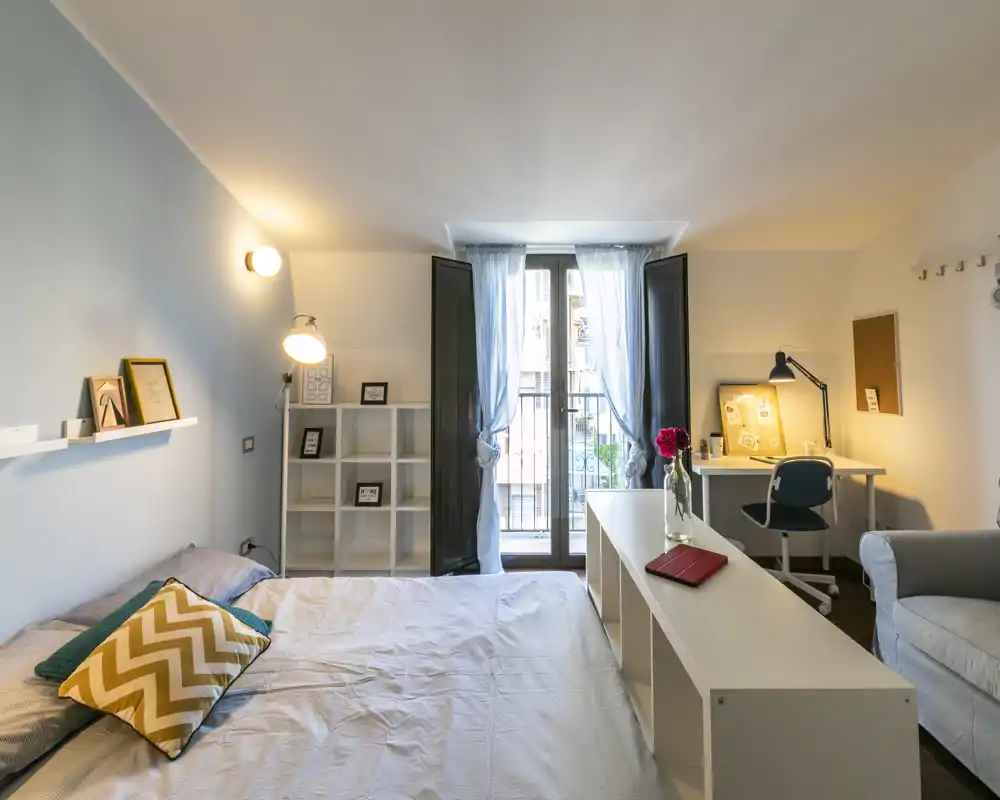 Dormitorio luminoso y con estilo, con cama doble, estantería, escritorio y balcón que aporta luz natural — ideal para estancias cómodas o para teletrabajar.