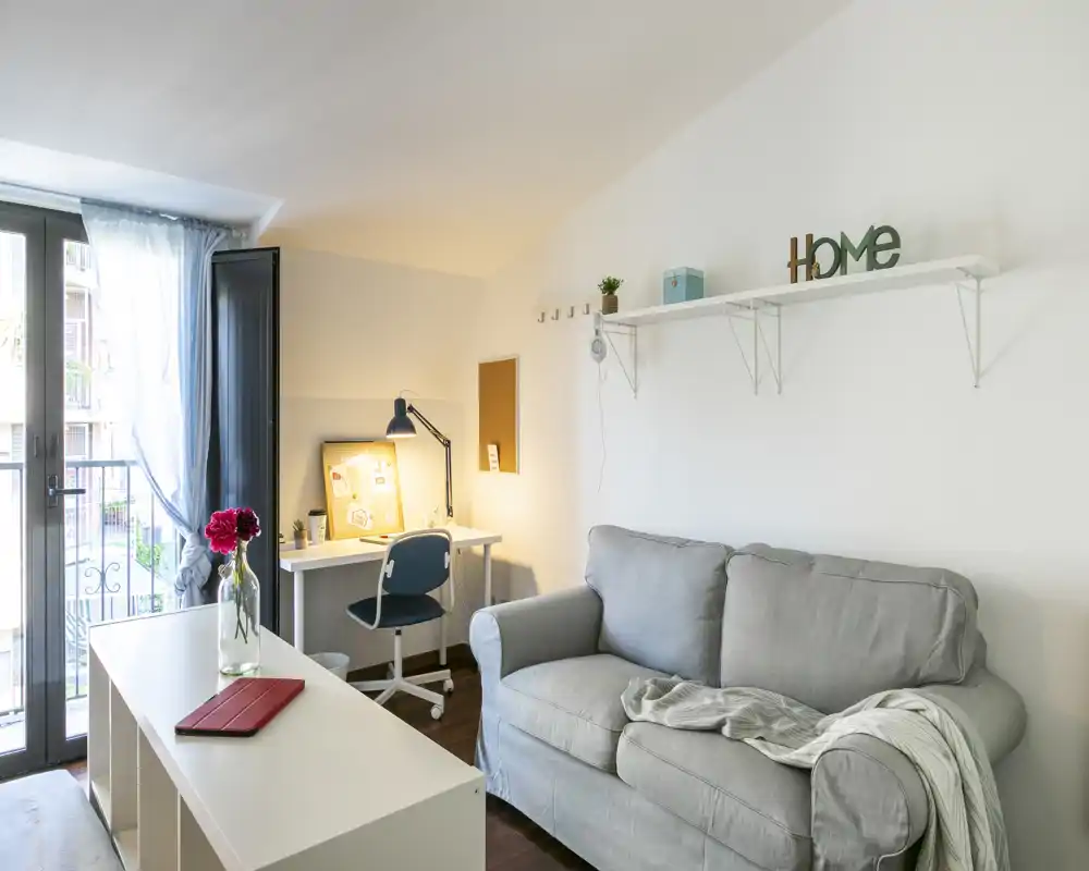 Salón luminoso y acogedor con un cómodo sofá gris, un escritorio pequeño y acceso al balcón que aporta luz natural — ideal para relajarse o teletrabajar.