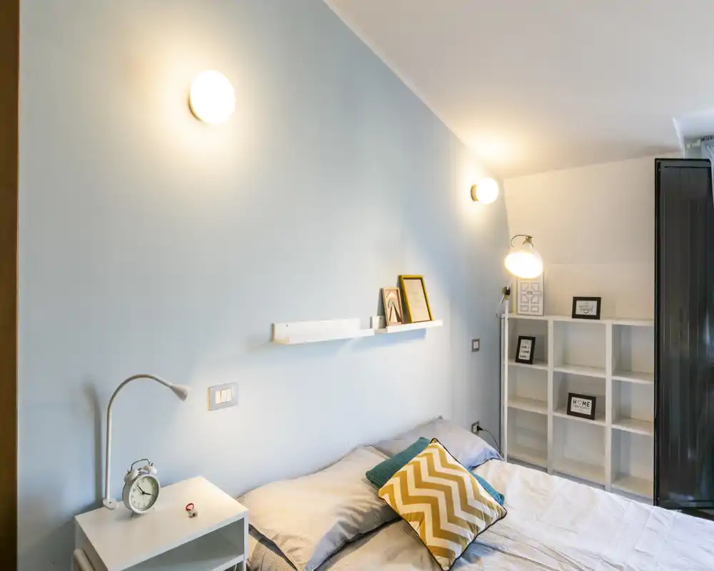 Dormitorio luminoso y acogedor con cama doble, cojines decorativos y mesita; iluminación de pared y estanterías crean un ambiente agradable.