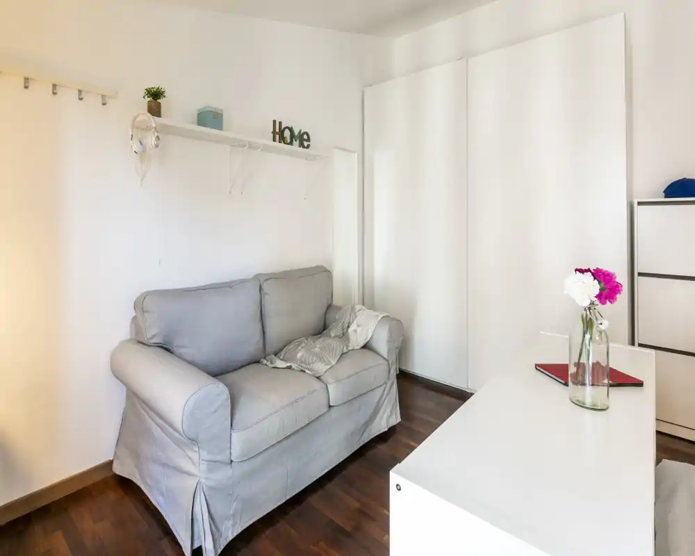 Pequeña sala luminosa con un sofá de dos plazas gris, estantería decorativa y muebles blancos; el suelo de madera y el estilo sencillo crean un ambiente acogedor.