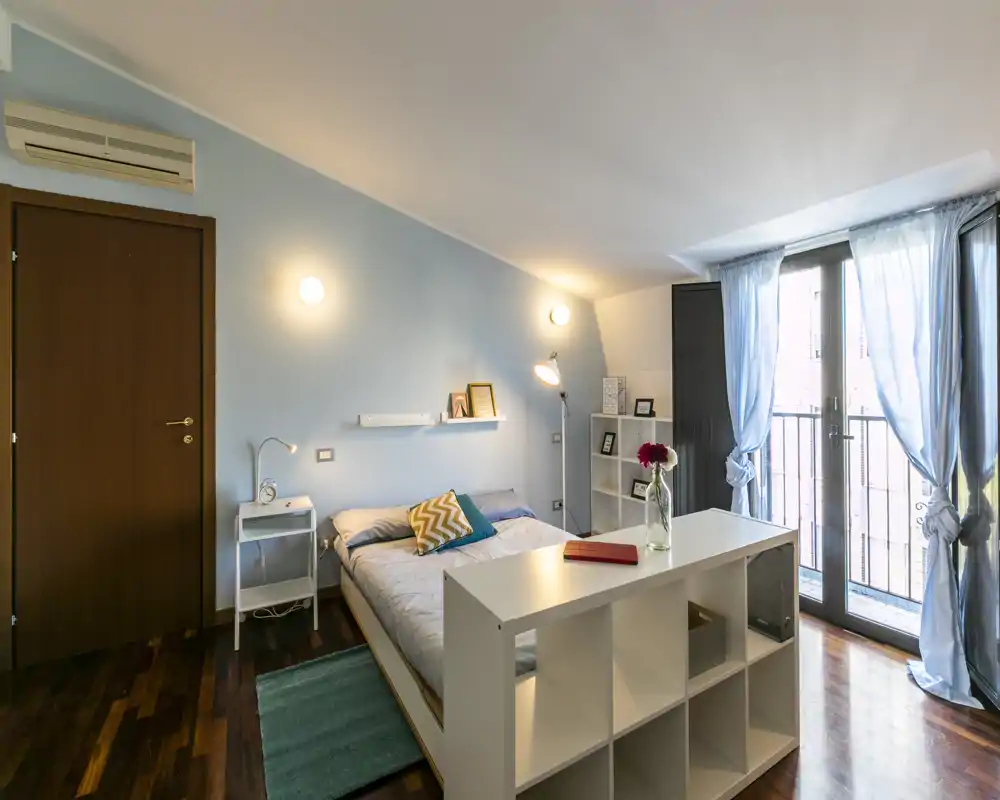 Dormitorio luminoso y bien decorado con cama doble, mesita, estantería modular y puertas al balcón que aportan luz natural.