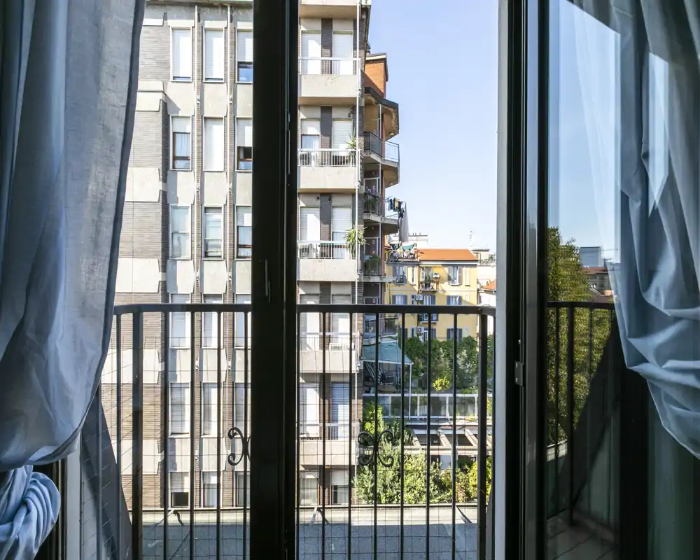 Balcón soleado enmarcado por cortinas translúcidas, con vistas a edificios residenciales y copas de árboles — ideal para mostrar un espacio exterior urbano.