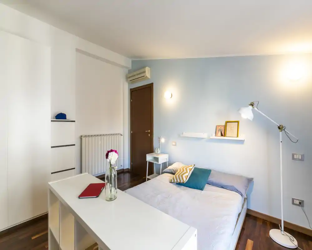 Dormitorio bien iluminado con cama doble, mesita, lámpara de pie y estanterías minimalistas. Pared azul suave y suelo de madera crean una sensación acogedora y moderna, ideal para alquileres temporales.