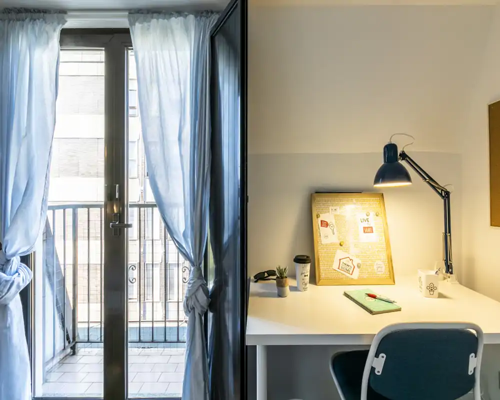 Rincón de salón luminoso con un escritorio junto a la puerta del balcón; la luz natural entra a través de cortinas finas y una lámpara de escritorio ilumina un espacio de trabajo ordenado con pequeños detalles decorativos.