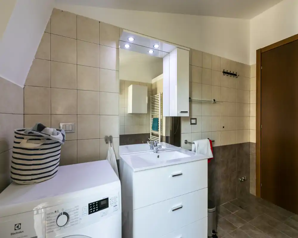 Baño moderno y limpio con mueble lavabo blanco, espejo grande con luces, paredes y suelo alicatados y una lavadora junto al lavabo.