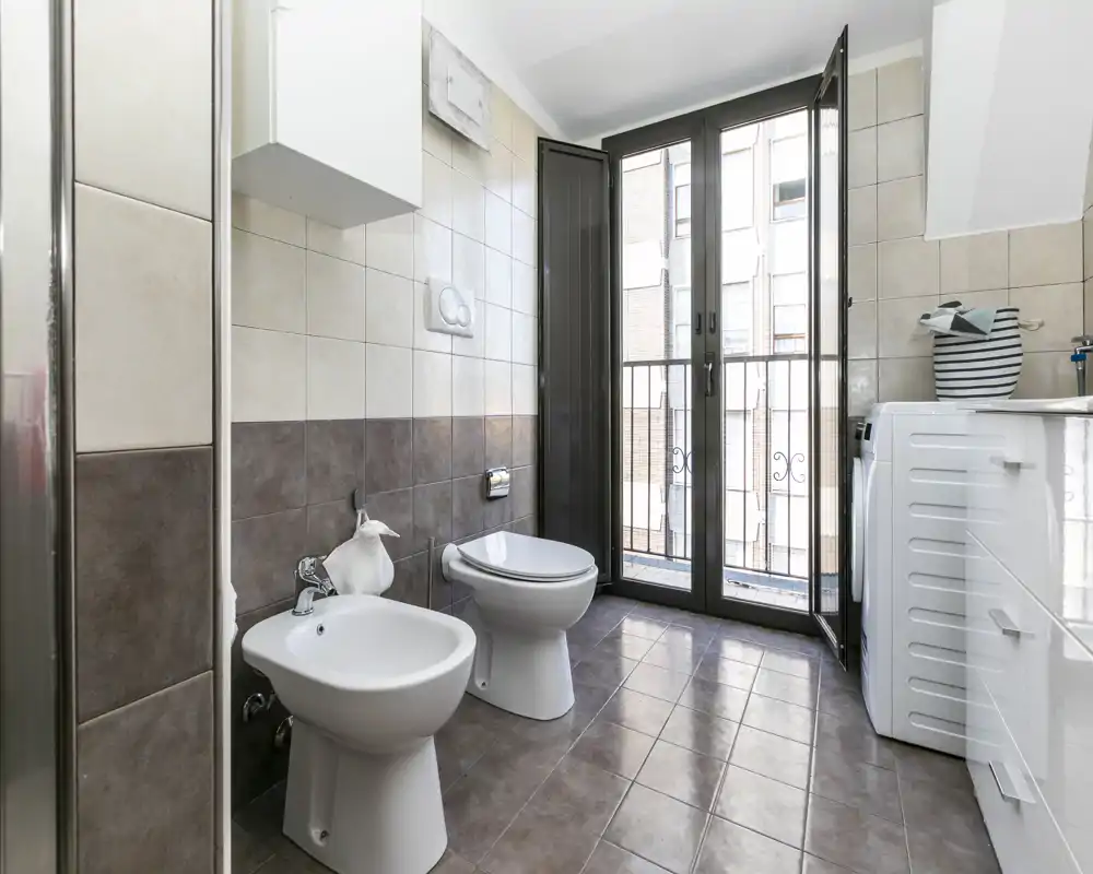 Baño moderno y luminoso con bidé y inodoro, paredes y suelo alicatados y una gran puerta de cristal que deja entrar luz natural.