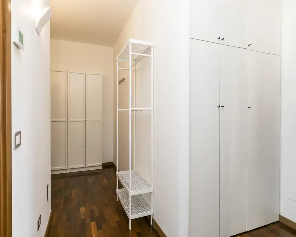 Entrada/pasillo minimalista con armarios empotrados blancos y una estantería independiente sobre suelo de madera cálida. Espacio luminoso y ordenado, ideal para abrigos y almacenamiento.