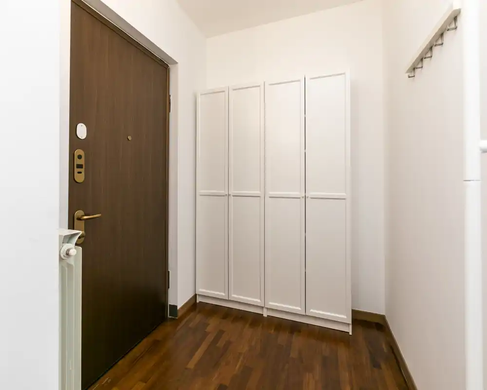 Entrada de apartamento luminosa y minimalista con puerta de madera, armario blanco y perchas en la pared sobre suelo de parquet cálido — primera impresión ordenada y acogedora.