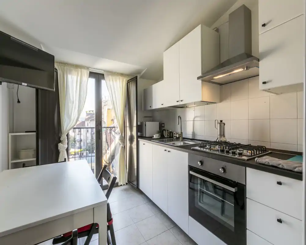 Cocina moderna y luminosa con muebles blancos, placa de gas y horno empotrado, además de una pequeña mesa y acceso al balcón que aporta luz natural.
