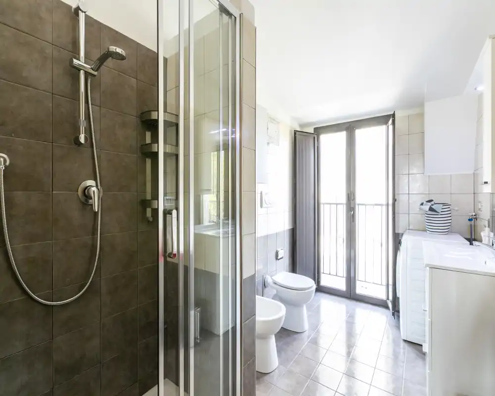 Baño moderno y luminoso con mampara de ducha de cristal, bidé y inodoro, lavabo con armario y una gran ventana/puerta que aporta mucha luz natural.