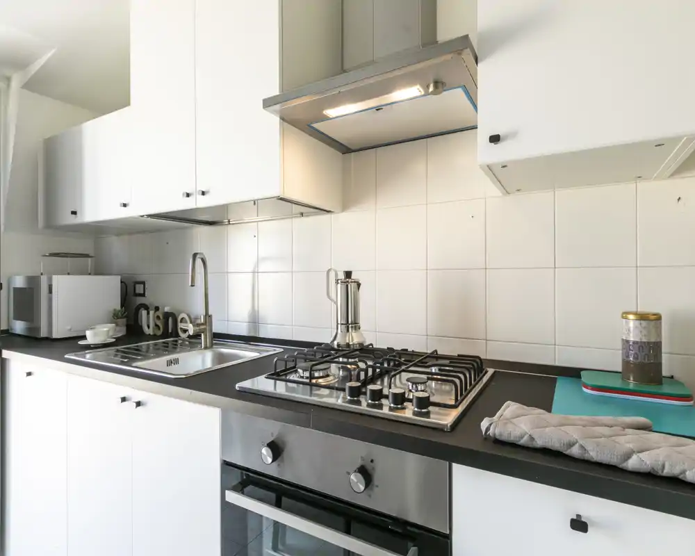 Cocina moderna y luminosa con armarios blancos, horno empotrado, placa de gas y fregadero de acero. Encimera ordenada con pequeños electrodomésticos y detalles acogedores.