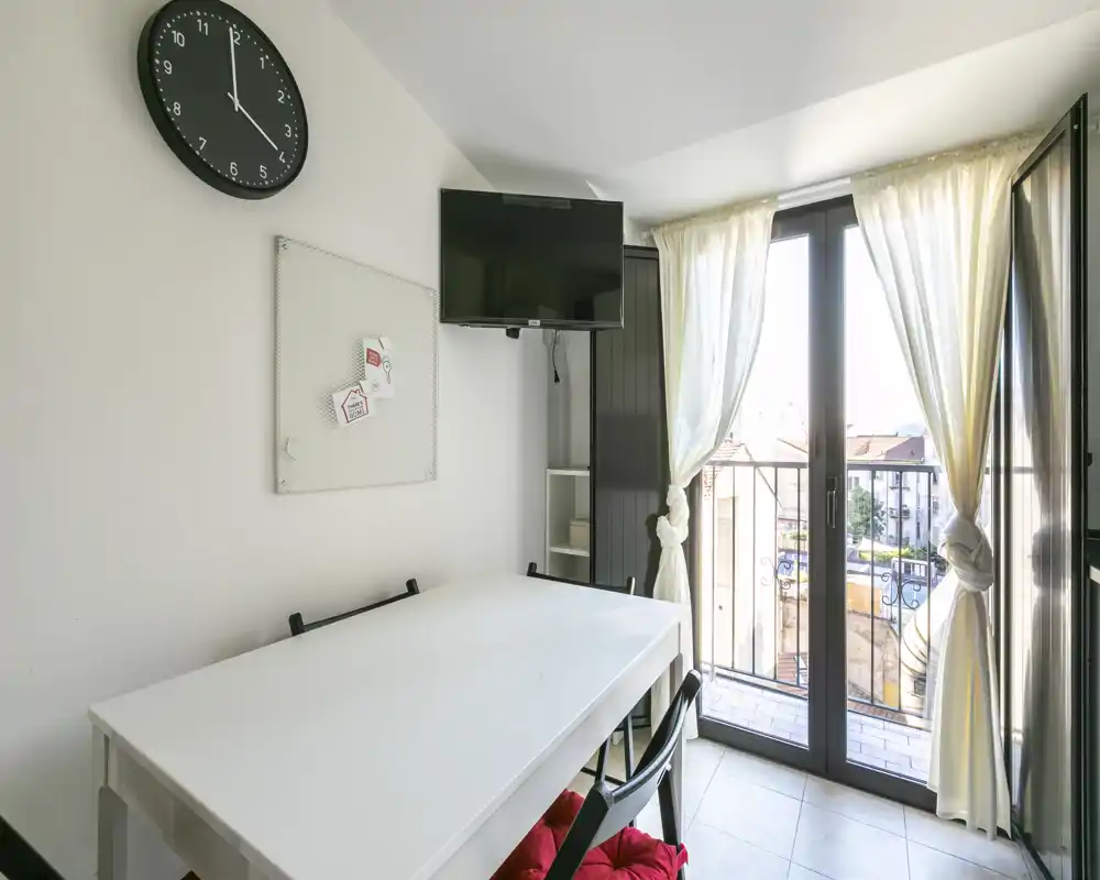 Zona de comedor luminosa y ordenada con mesa blanca, sillas con cojines y grandes puertas de cristal que dan a un pequeño balcón — ideal para comer con luz natural.
