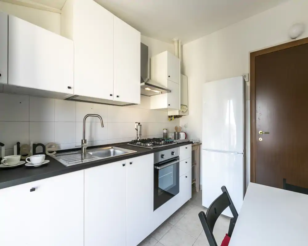 Cocina moderna y luminosa con muebles blancos, horno integrado y placa de gas, fregadero de acero inoxidable y frigorífico independiente; mesa pequeña y sillas para un apartamento urbano compacto.