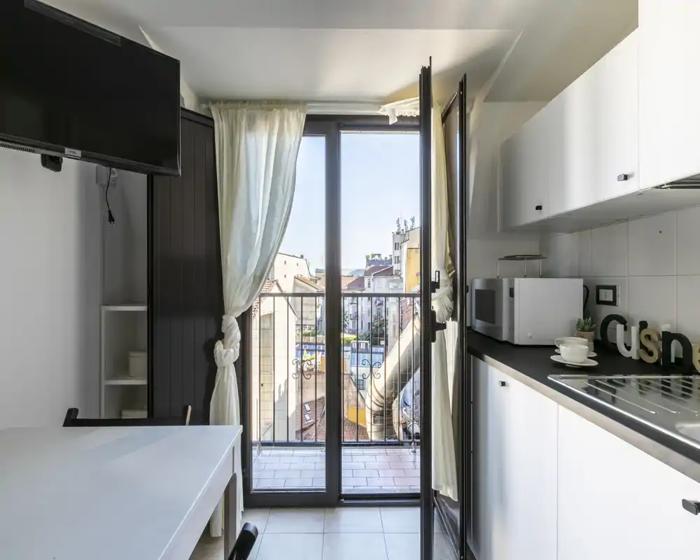 Cocina compacta con muebles blancos, microondas y fregadero, que se abre a un pequeño balcón con vistas a la ciudad. Buena luz natural y mobiliario moderno y sencillo, funcional para estancias cortas.