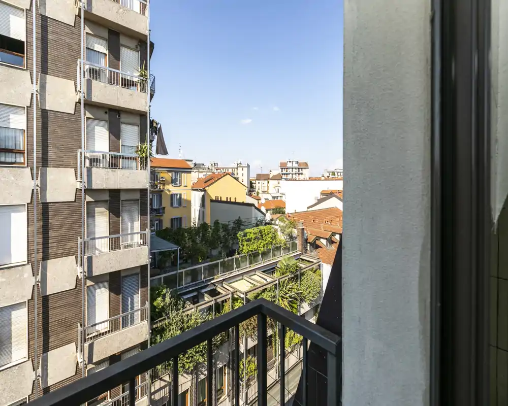 Balcón soleado con barandilla de metal con vistas a edificios y terrazas ajardinadas; ideal para tomar el café por la mañana con cielo despejado.