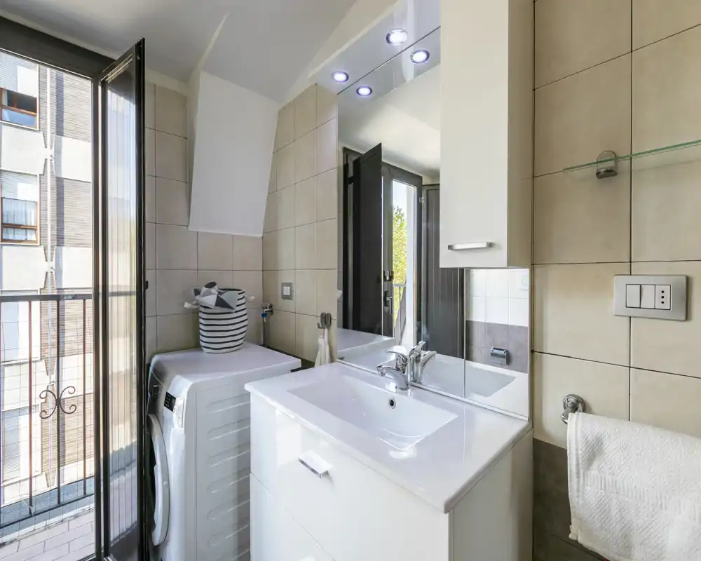 Baño moderno con lavabo blanco, espejo con luces integradas, toallero y lavadora junto a la puerta del balcón abierta que deja entrar luz natural.