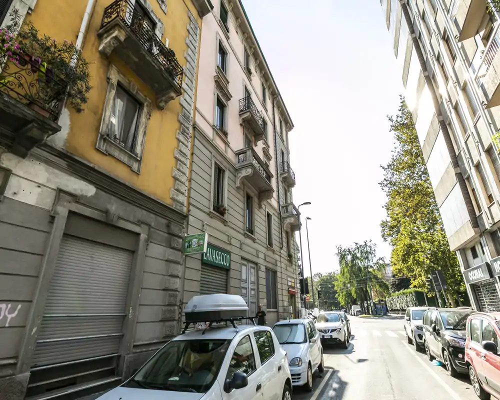 Exterior a nivel de calle con coches aparcados, edificios residenciales y acera arbolada; luz diurna brillante y fachada urbana bien definida.